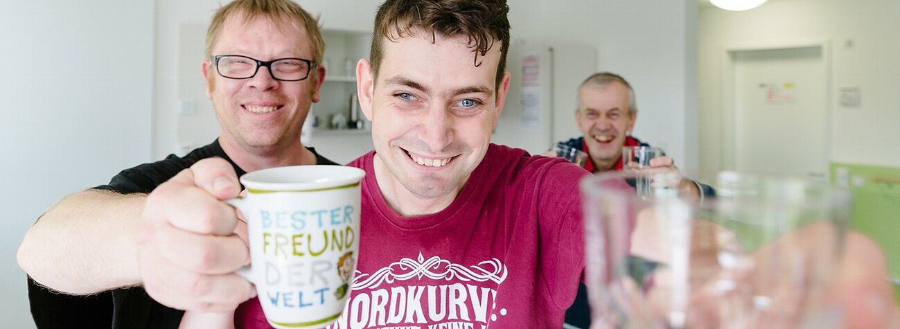 Bewohner des Wohnheims in Gunzenhausen sind gute Freunde - drei Männer prosten dem Fotografen zu - einer davon mit einer Tasse mit der Aufschrift "Bester Freund der Welt"