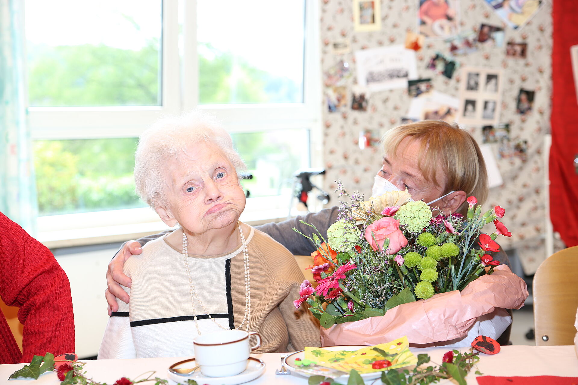 Eine alte Dame sitzt an einem Tisch. Auf dem Tisch steht ein Blumenstrauß und eine Tasse..